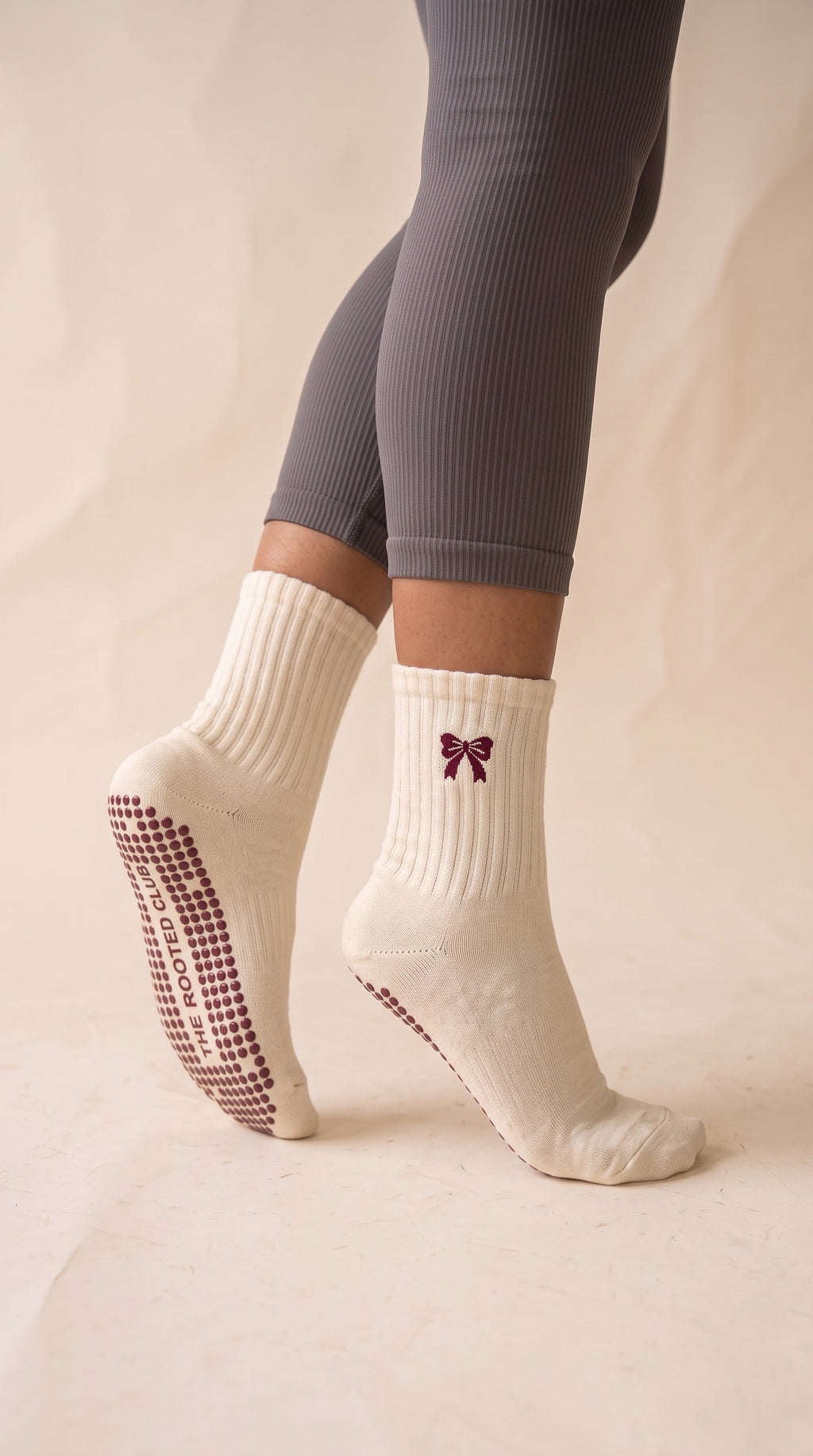 A person wearing ribbed cream socks with grip dots on the sole and a small burgundy bow graphic on the side, paired with gray ribbed leggings against a warm beige backdrop.